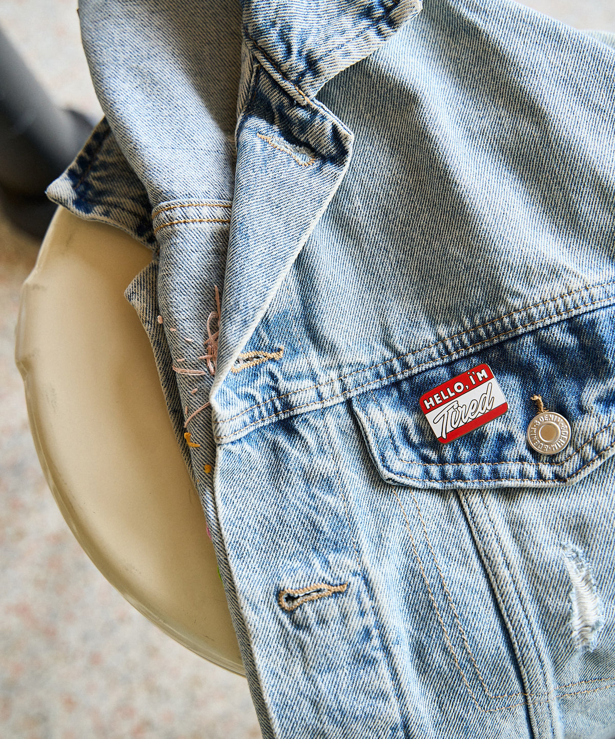 Hello, I'm Tired enamel pin worn on the chest pocket of a light-wash denim jacket