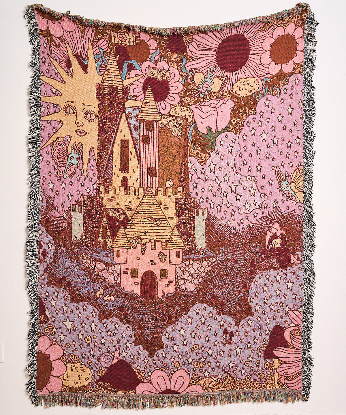 Autumnal Wood woven blanket hanging on a wall, showing a pink castle among clouds and flowers under a smiling sun