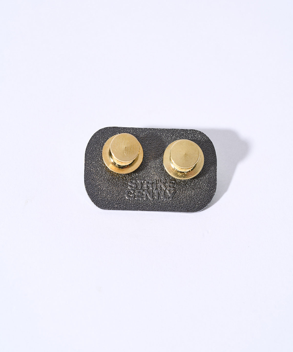 Back of the Buy a Man Eat Fish pin showing the black backing, two gold clutches and Strike Gently backstamp