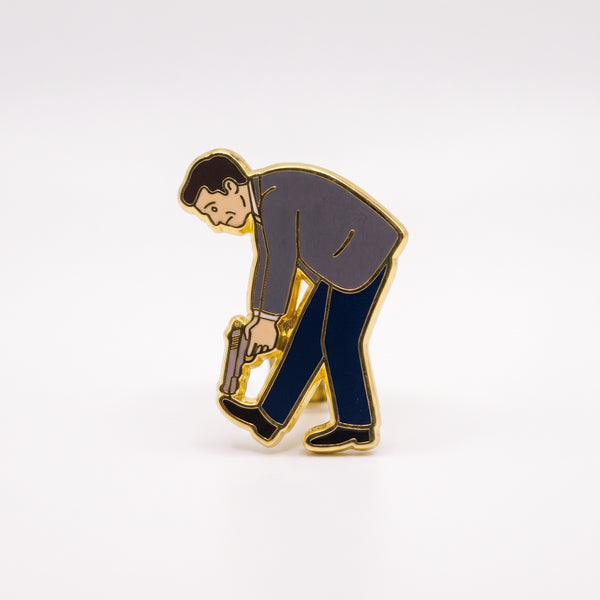 Shot In The Foot enamel pin, a man in a gray jacket and navy pants bending over to aim a gun at his own foot