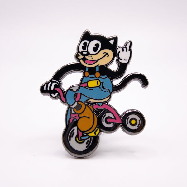 Cat Riding Tricycle enamel pin, a grinning black cartoon cat in blue overalls flipping the bird while riding a pink tricycle