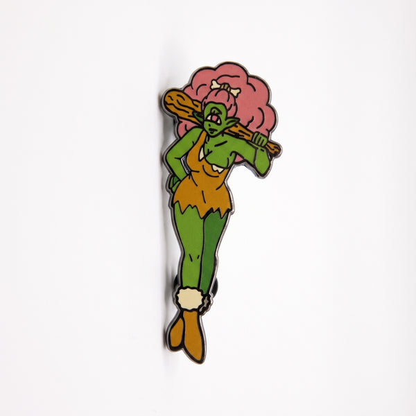Caveman Mermaid enamel pin, a green one-eyed cavewoman with pink hair and a bone, posing in a tan fur dress with a club