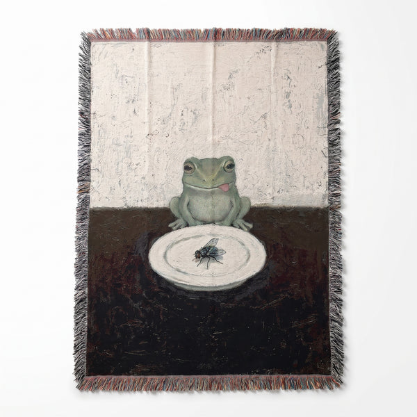 Frog And Fly woven blanket showing a green frog licking its lips at a dark table over a single fly served on a white plate