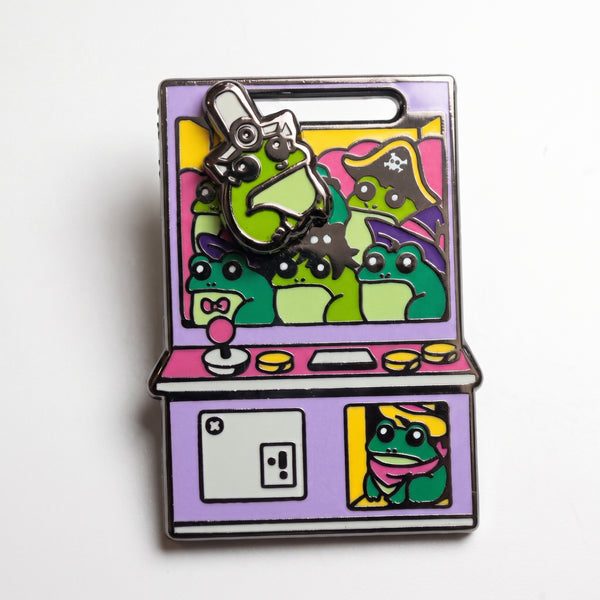 Frog Arcade Claw Machine enamel pin, a purple arcade claw machine packed with green frogs, one in a pirate hat, with the claw grabbing a frog