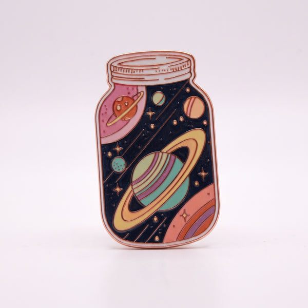 Galaxy In A Jar enamel pin, a glass mason jar holding a navy night sky with a pastel ringed planet, stars and comets