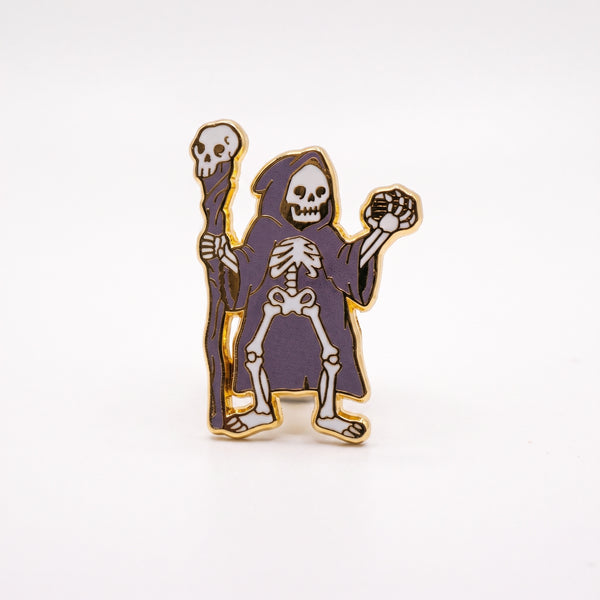 Grim Reaper enamel pin, a skeleton in a hooded purple robe holding a skull-topped staff and raising a bony hand