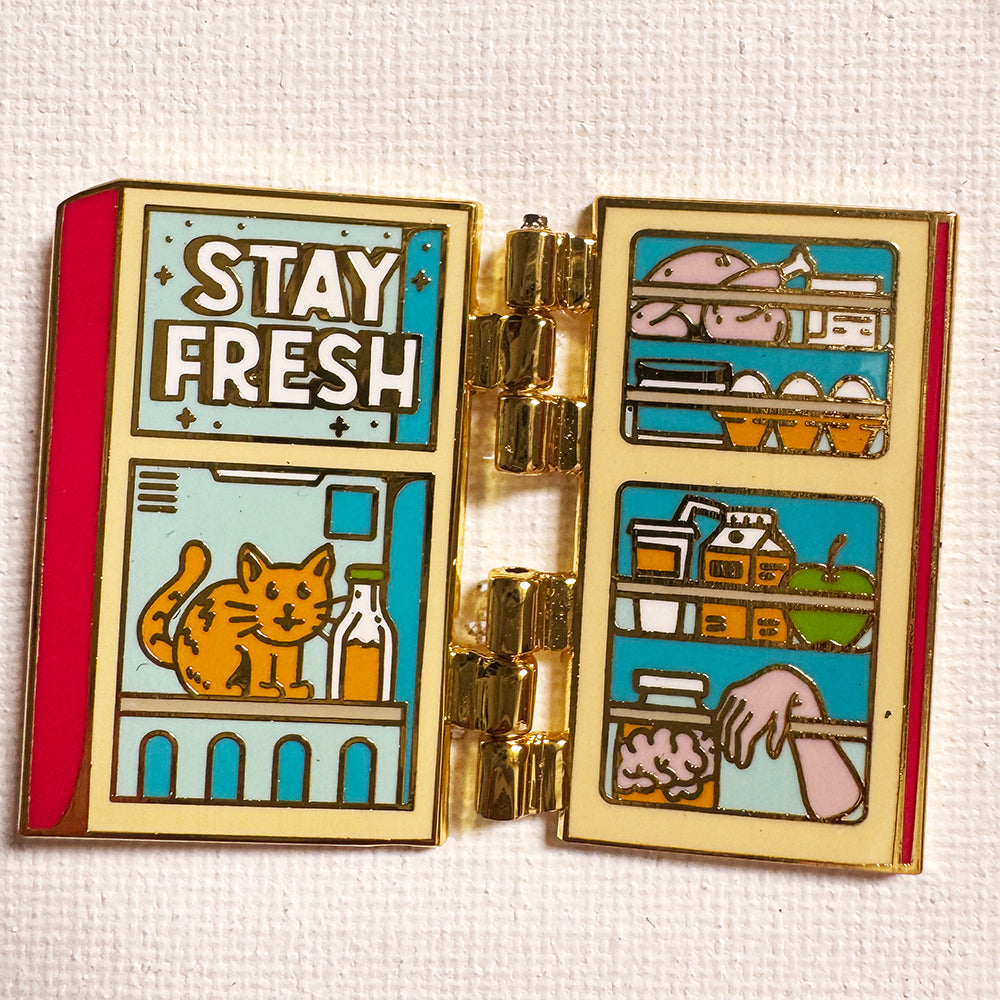 Fridge Deluxe enamel pin opened on gold hinges, showing shelves of food and a cat inside, on a white textured background