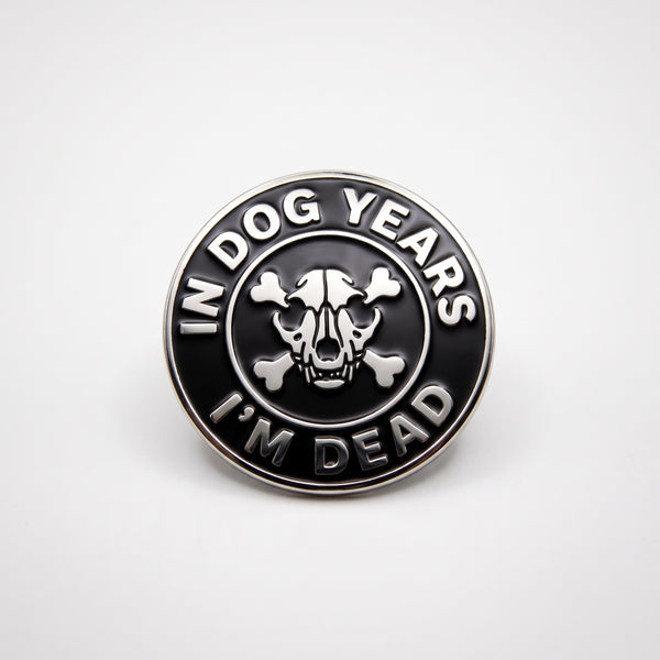 In Dog Years I'm Dead enamel pin, a round black badge with a silver dog skull and crossbones