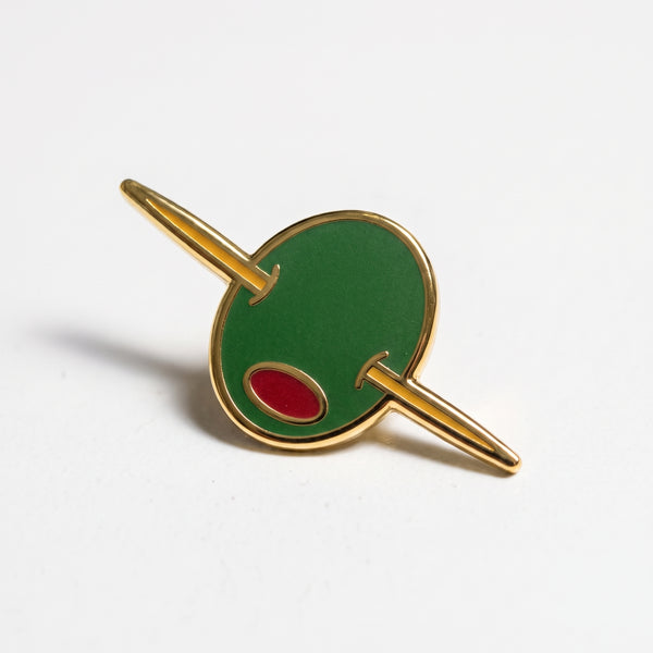 Olive Cocktail enamel pin, a green martini olive with a red pimento skewered on a gold cocktail pick