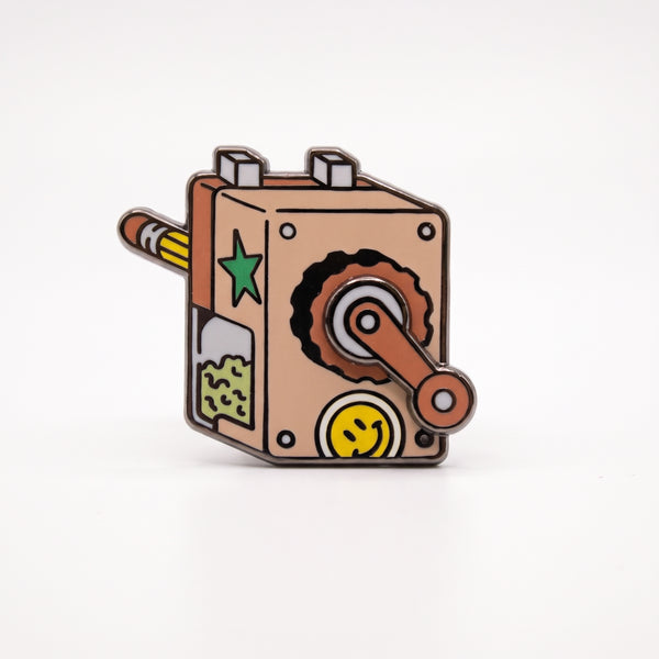Pencil Sharpener enamel pin, a tan crank pencil sharpener with a pencil, a smiley face sticker and a turning crank