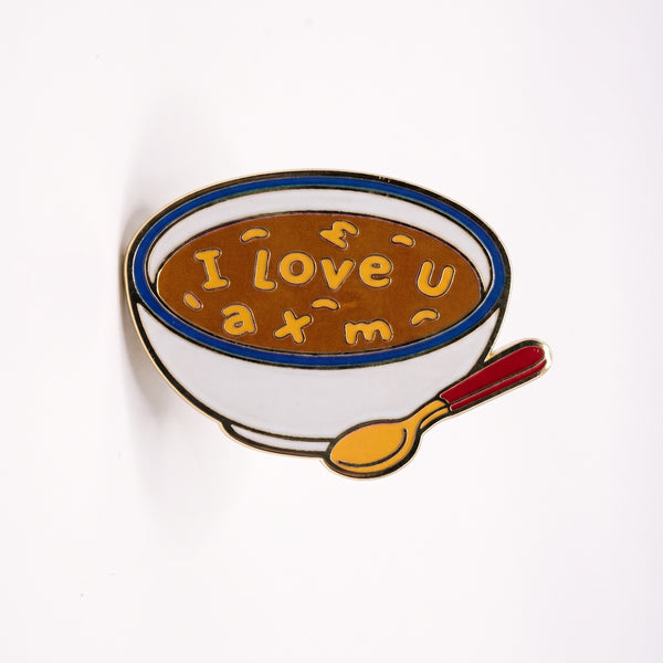 Soup Bowl enamel pin, a white bowl of brown alphabet soup spelling I love u with a gold spoon