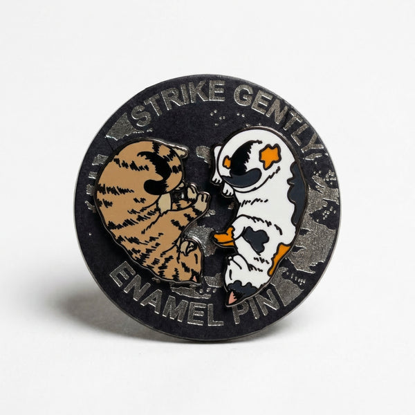 Round enamel pin with 'Strike Gently Enamel Pin' text and a spinning center showing two cats.