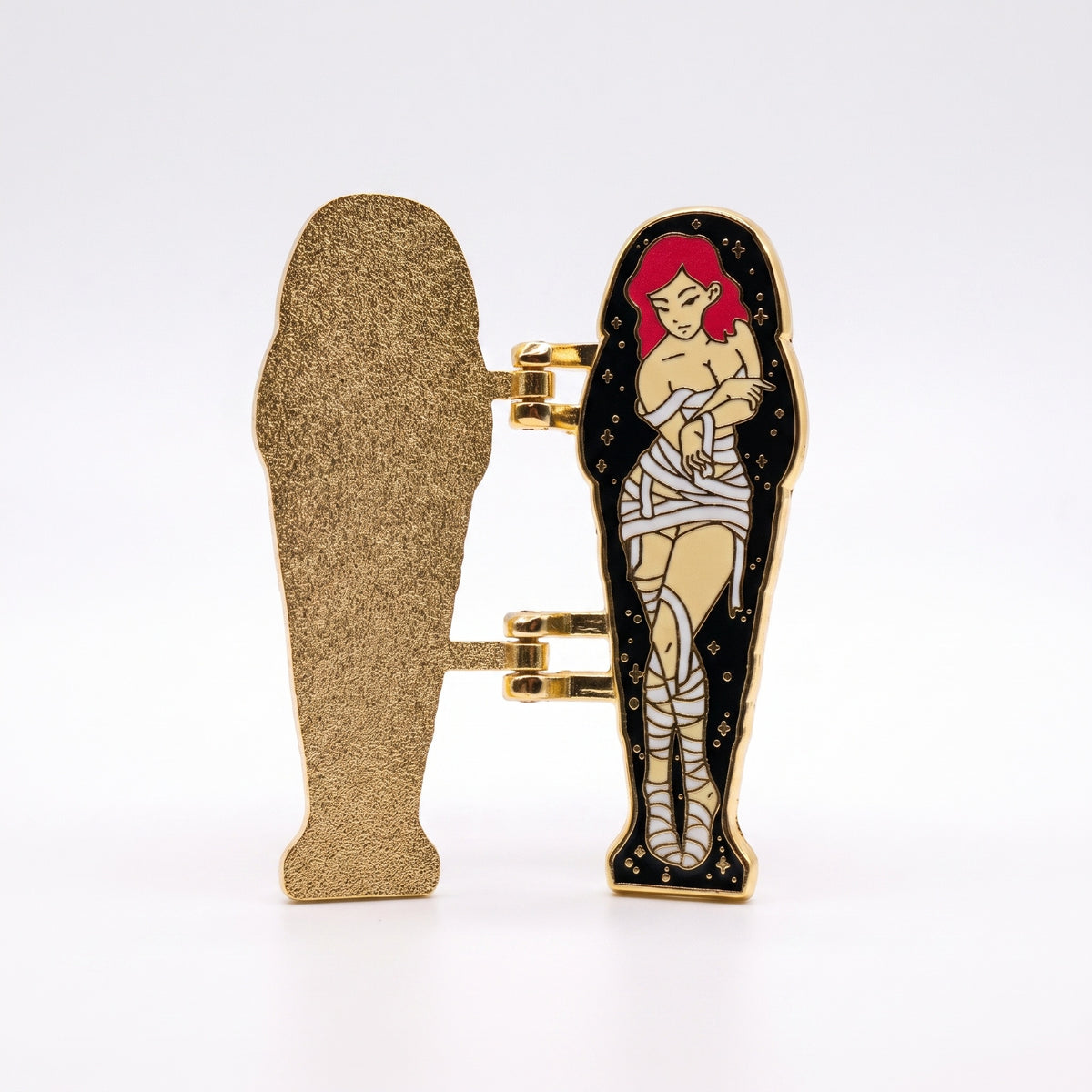 Sarcophagus enamel pin opened, showing a gold glitter lid and a red-haired mummy pin-up inside