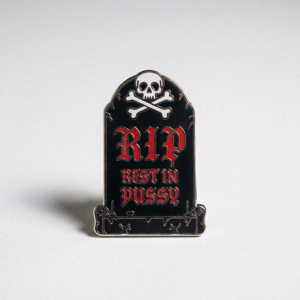 A black tombstone enamel pin with a skull and crossbones and red text reading RIP Rest in Pussy.