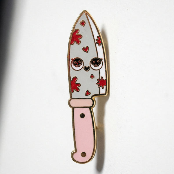 Enamel pin of a chef knife with a face, skull eyes, pink handle, and red blood splatters