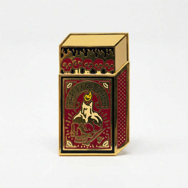 Gold and red enamel pin shaped like a matchbox with skulls and safety matches text