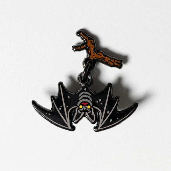 A two-piece enamel pin of a black bat dangling from a brown branch.